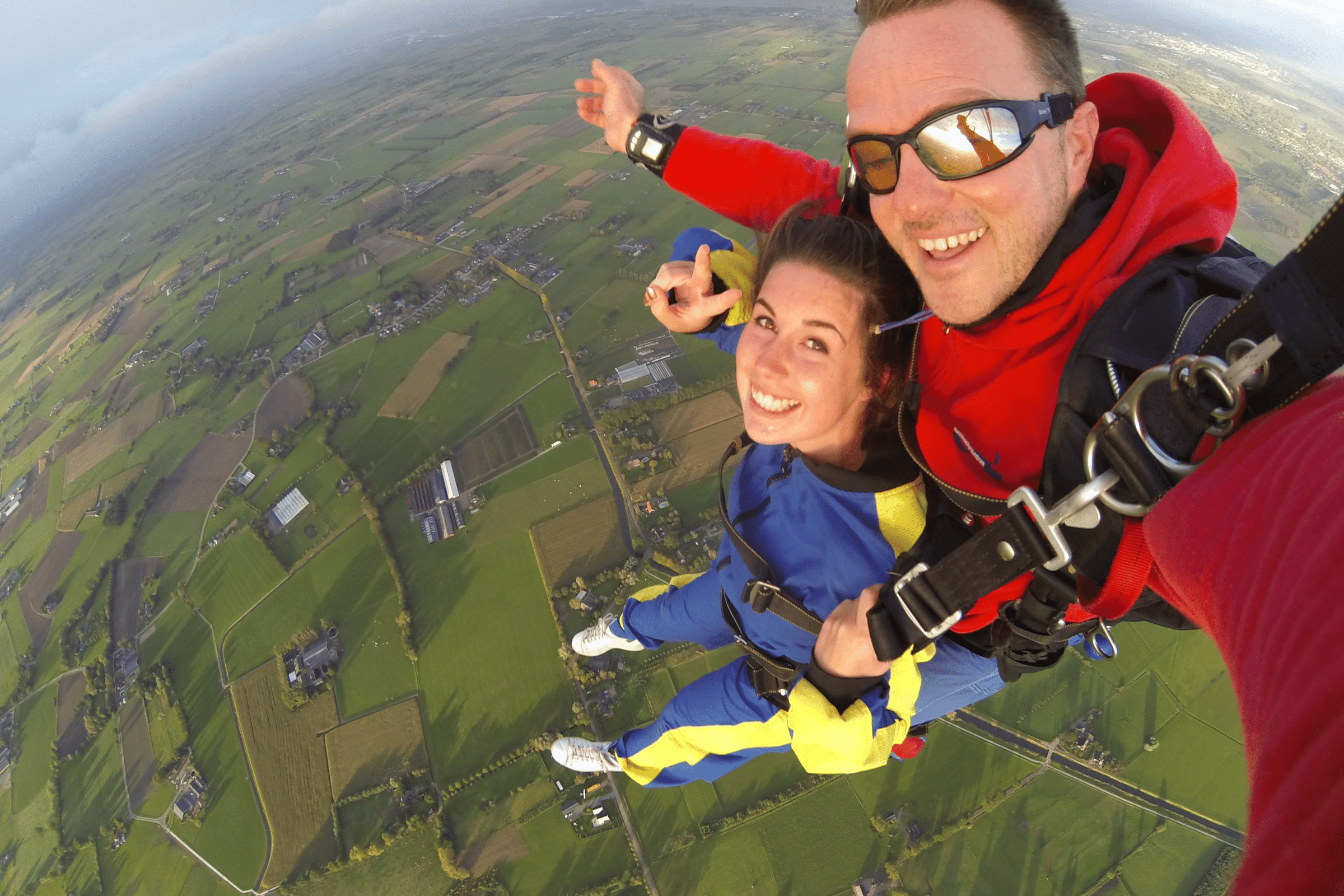 A white woman in her 30s and a white man in his 40s are doing a tandem skydive. They are high in the air above patchwork fields. They are both smiling.