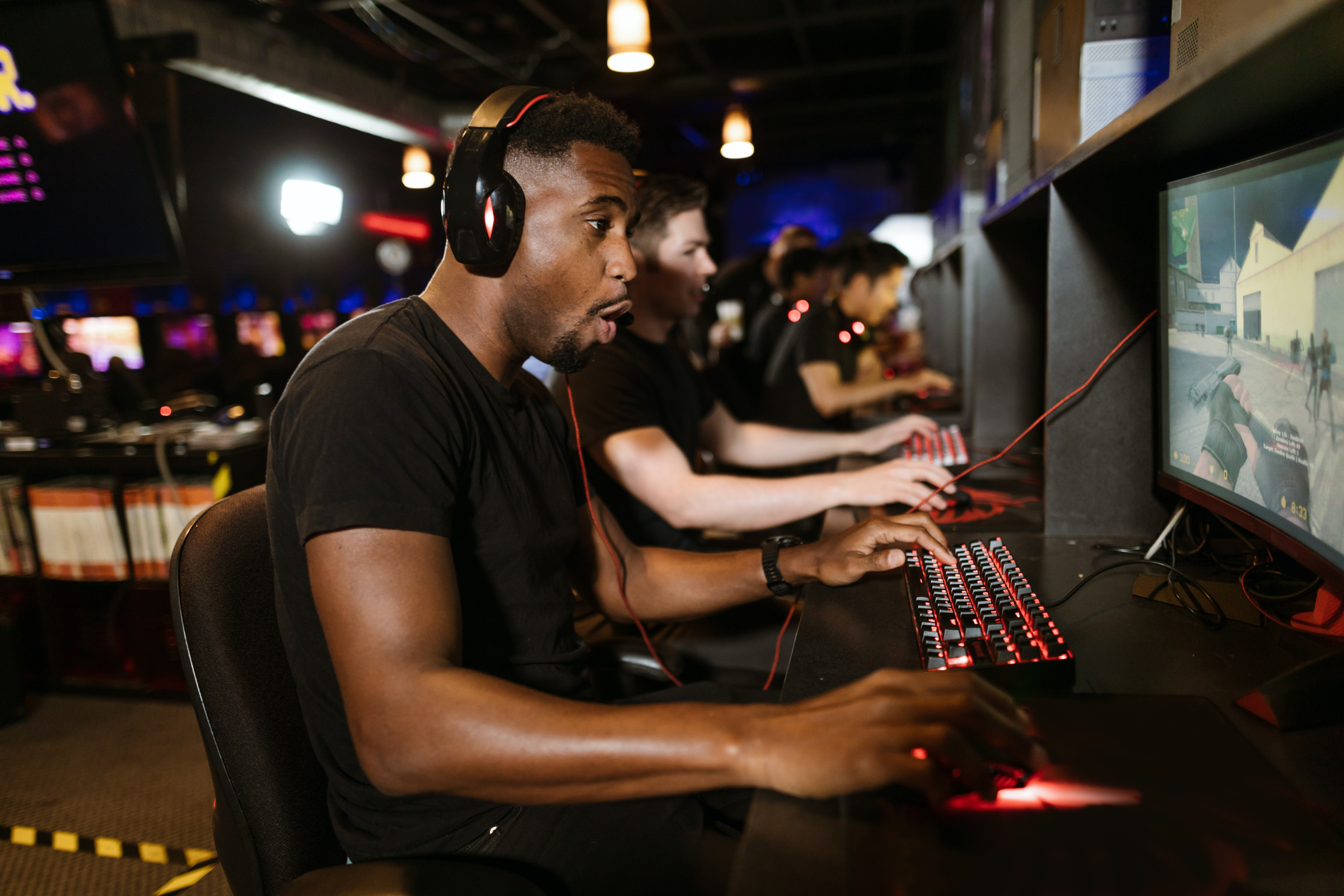 A team of gamers all playing computer games in a row. In the forground is a black man in his 20s, wearing a black t shirt and headphones. He has an excited and focussed expression