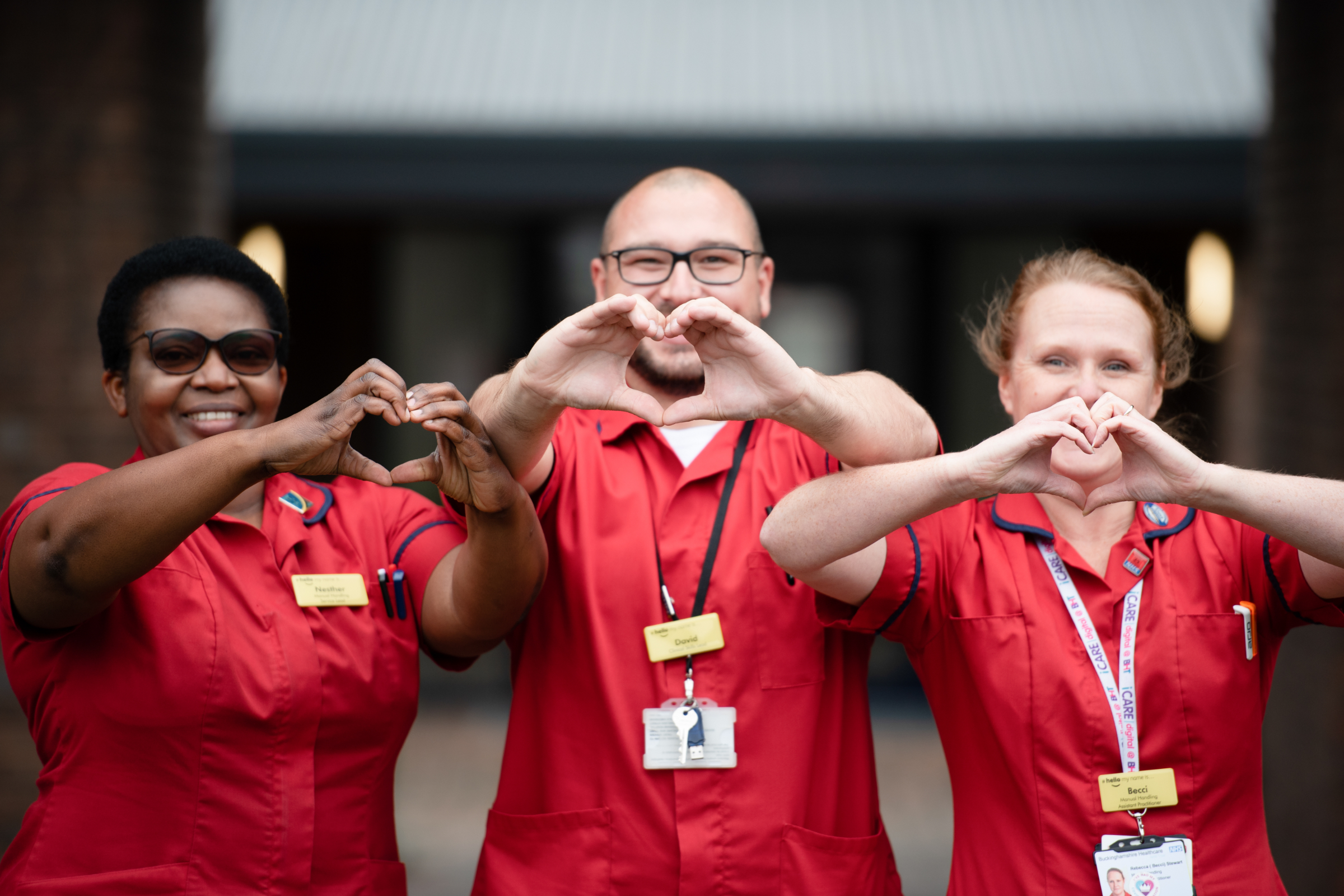 Three members of staff at SToke Mandeville Hospital; a black woman, a white ma wearing glasses and a white woman. They are all wearing red scrubs and making a heart shape with their hands.