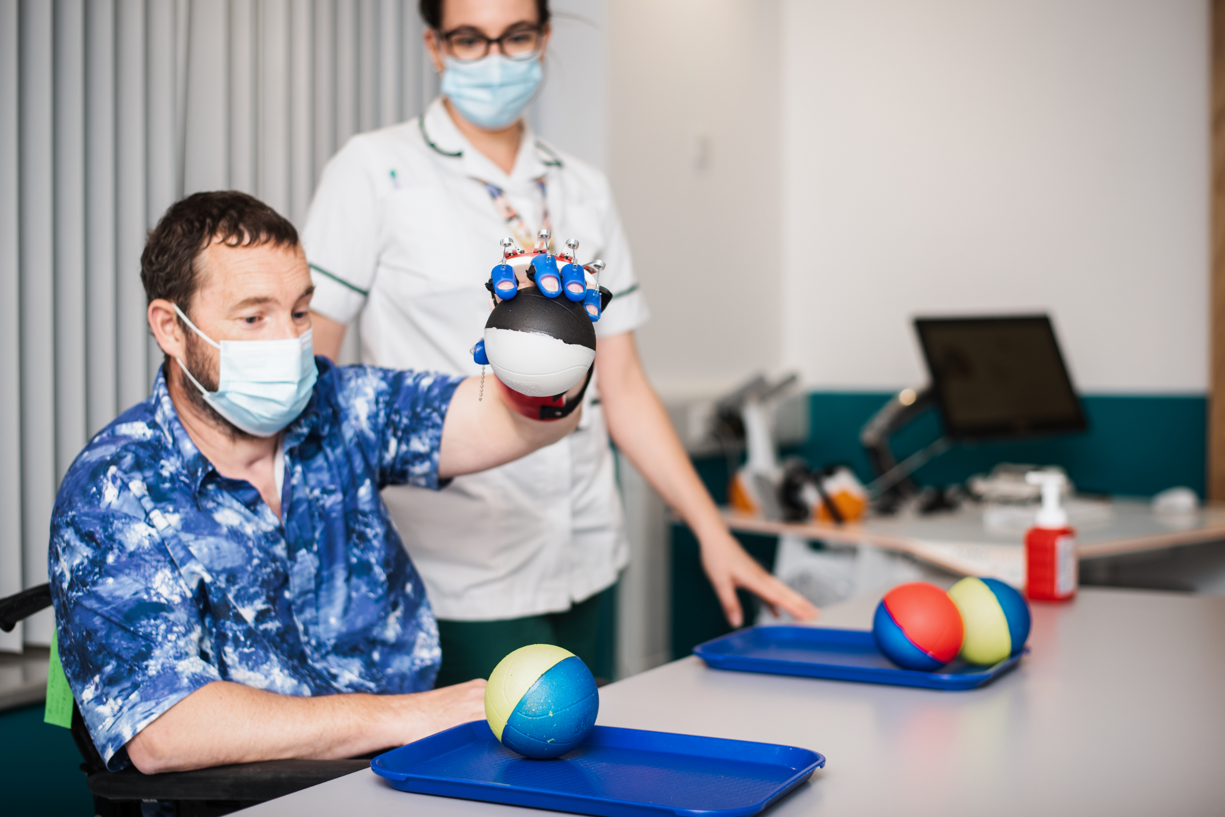 A spinal injury patient, a white man in his 40s, is holding a ball using a robotic arm. He's wearing a blu pattenred shirt and is using a wheelchair. A healthcare assistant is supporting him.