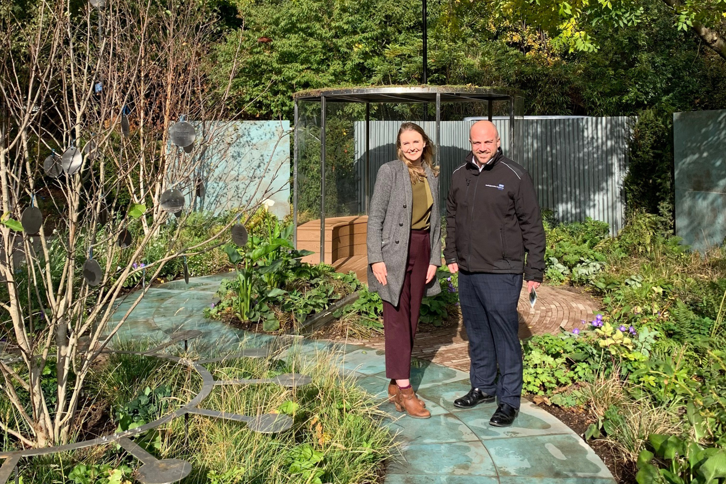 A white woman in her 30s and a white man in his 40s are stood in the new garden in winter coats. The copper-green path winds through the planting and a tree in the foreground has messages of thanks hanging in the branches