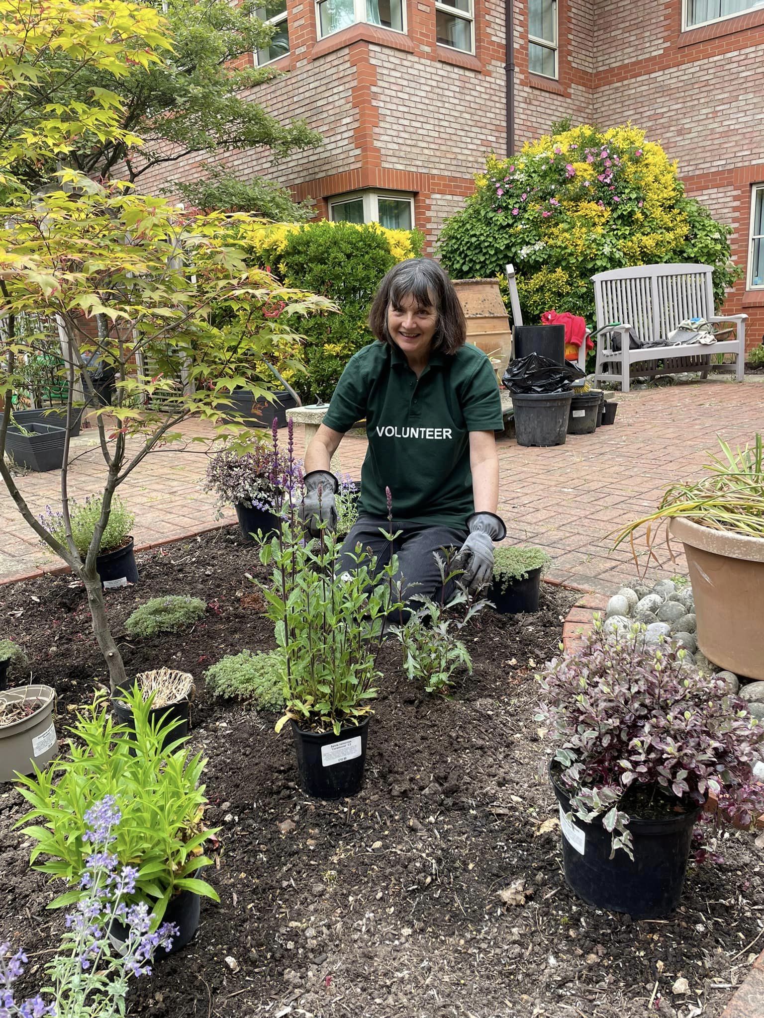 A female volunteer in a green tabard, kneeling next to a flower bed with pots of plants.