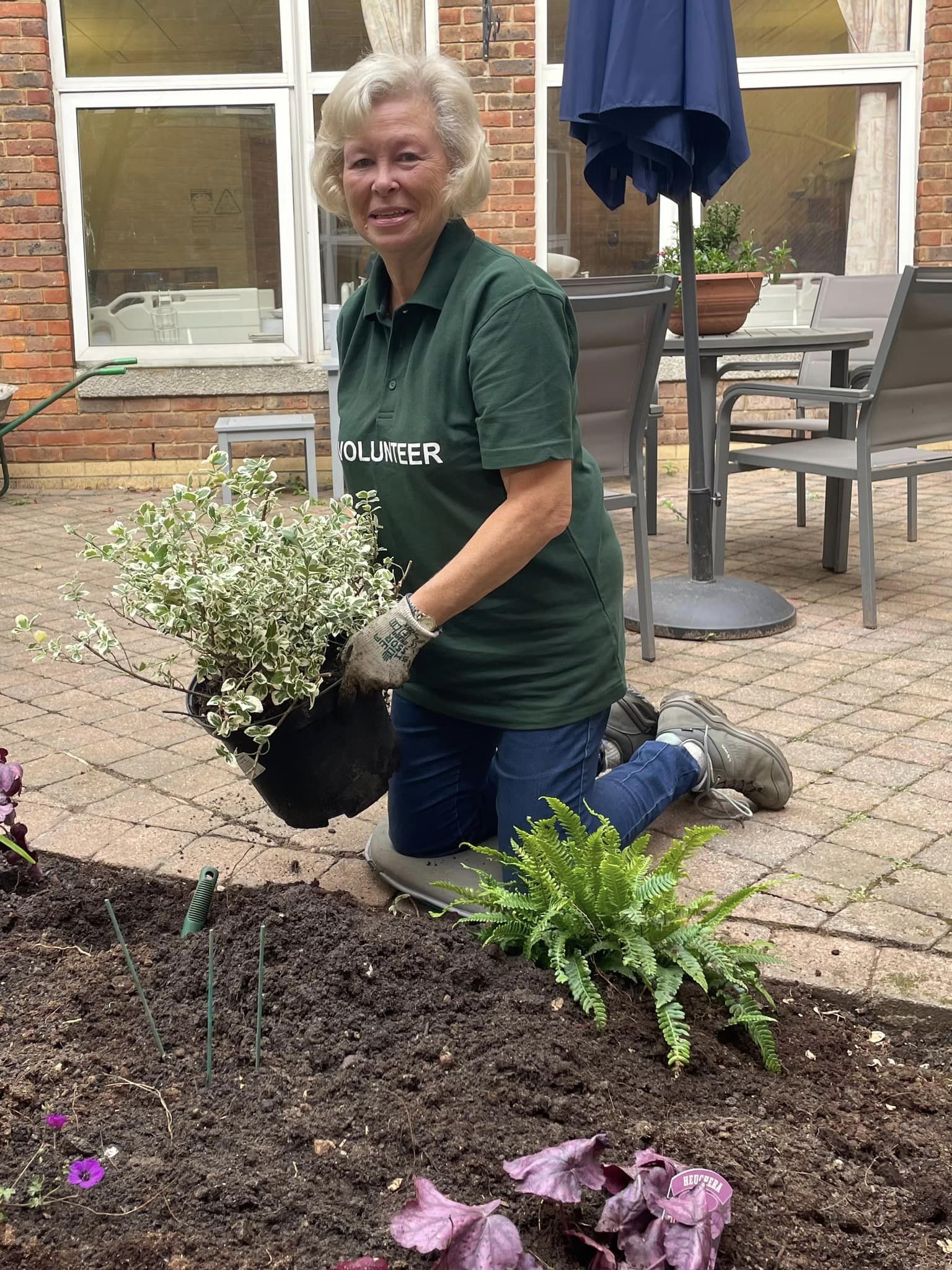 A female volunteer in a green tabard, kneeling next to a flower bed, holding a potted plant.