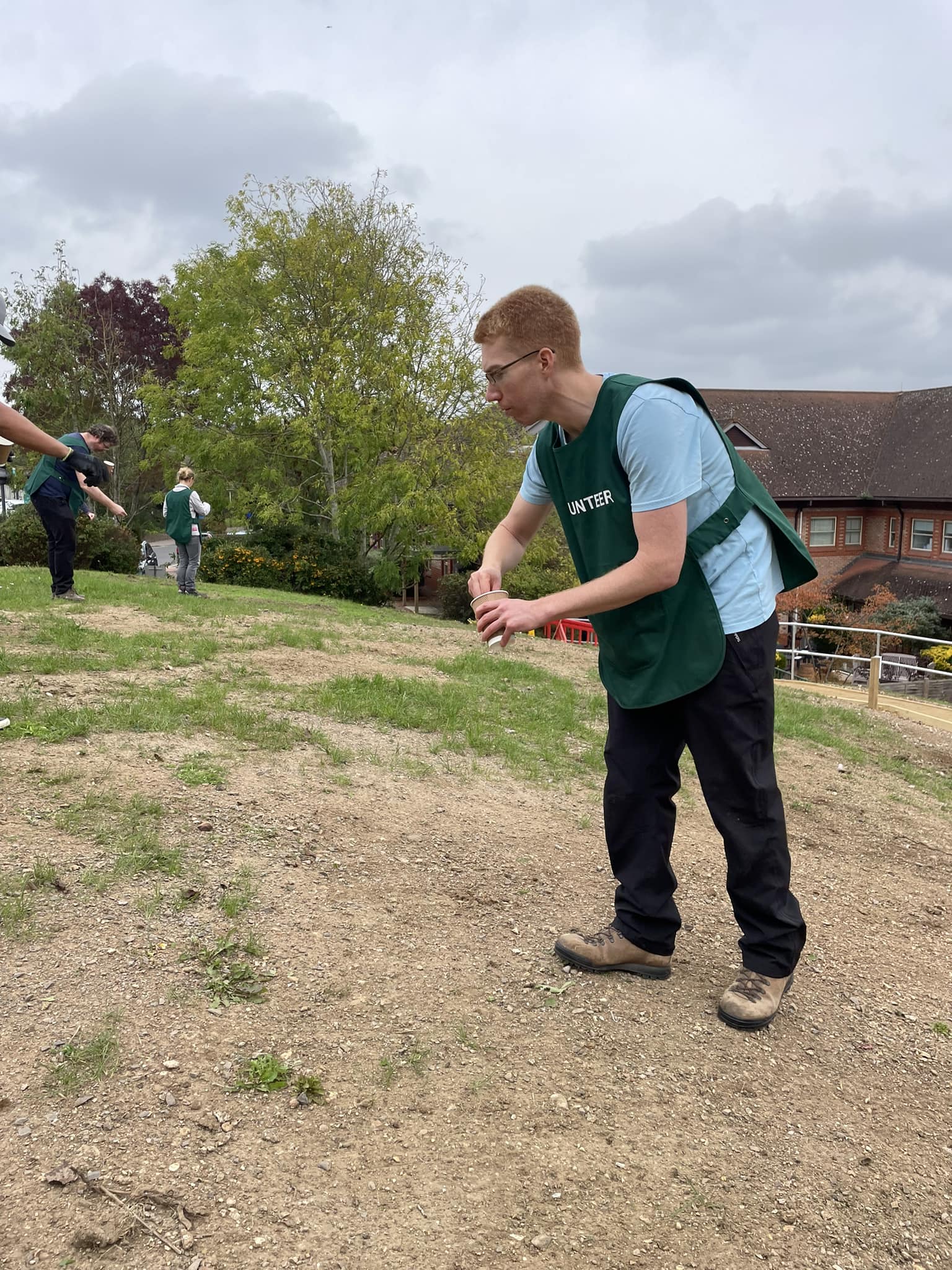 A volunteer in a green tabard spreading seeds over a lawn. More volunteers can been seen in the background doing the same.
