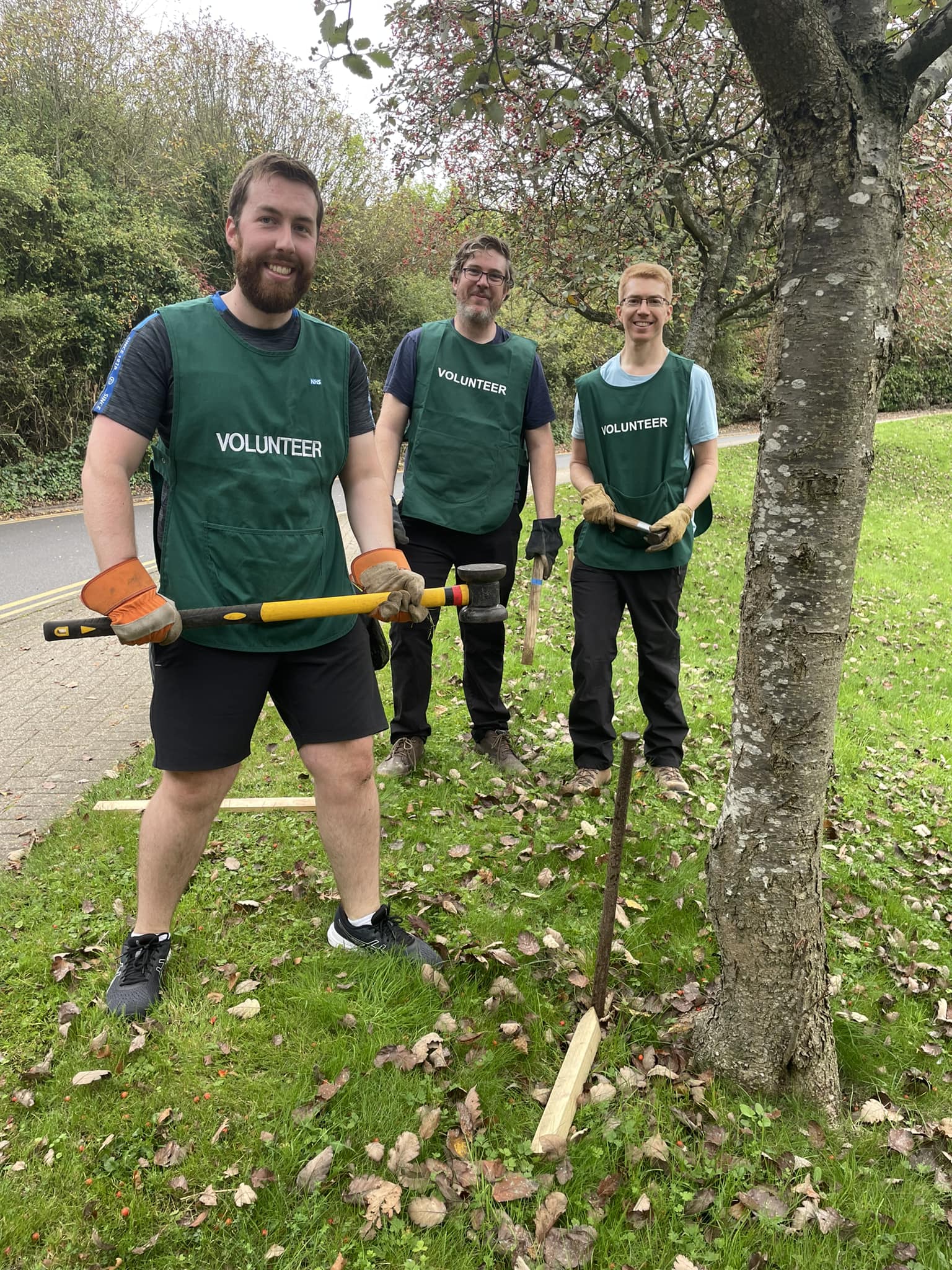 Three volunteers in green tabards, one holds a mallet and is standing next to a metal stake which has been driven into the ground.