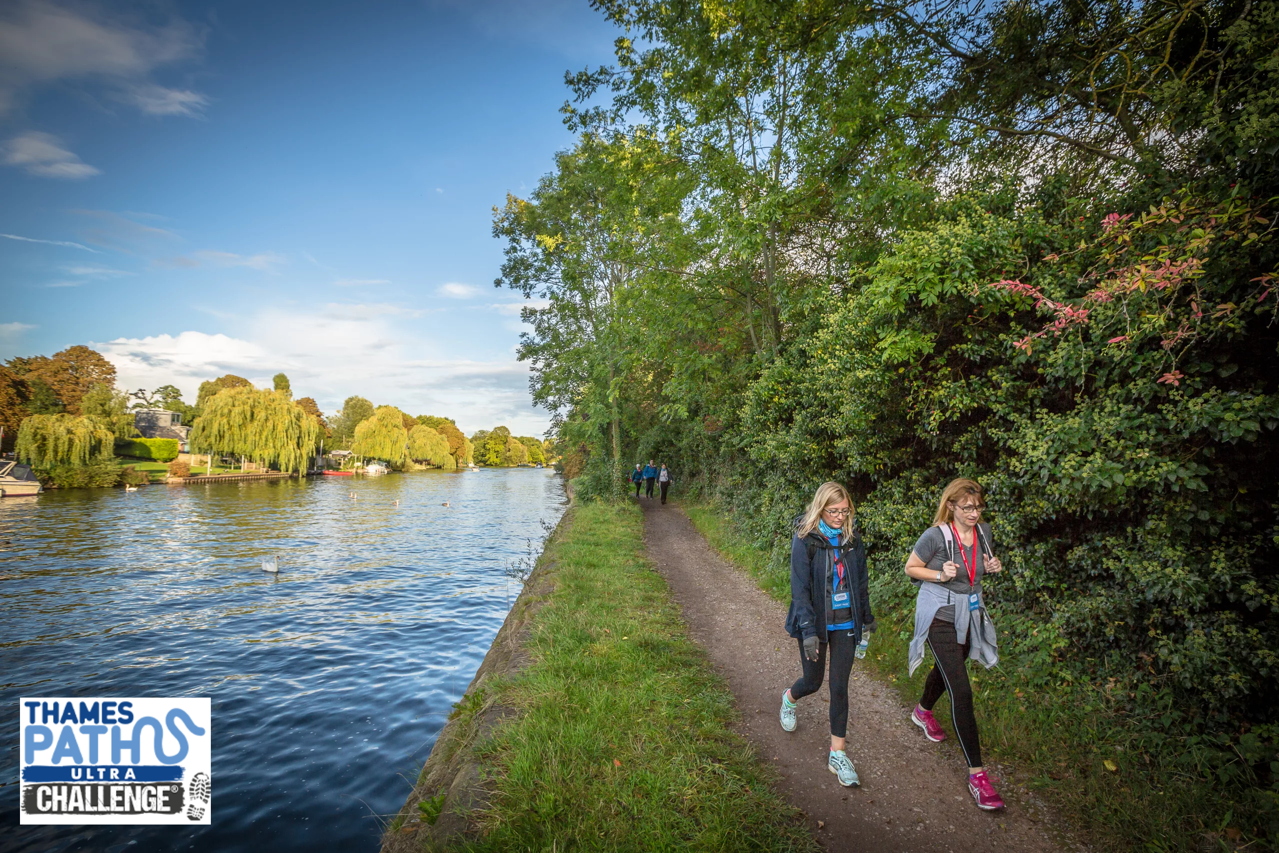 Two people taking on the Thames Path Challenge, walking along a towpath in the countryside.
