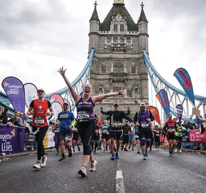 Marathon runner crossing Tower Bridge, in the foreground a runner wearing a purple Buckinghamshire Healthcare NHS Trust Charitable Fund running vest.
